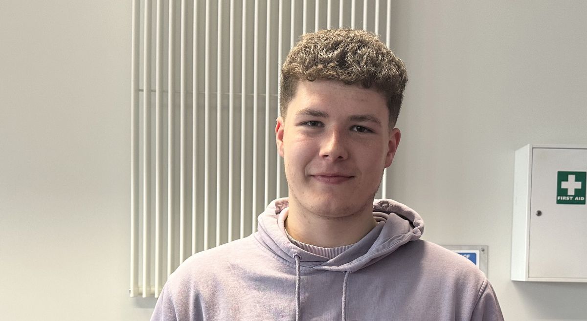 Jamie Rusk - young man with short curly hair, wearing a grey hoodie, smiling to camera pictured inside a classroom with wall mounted radiator and first aid kit in background.